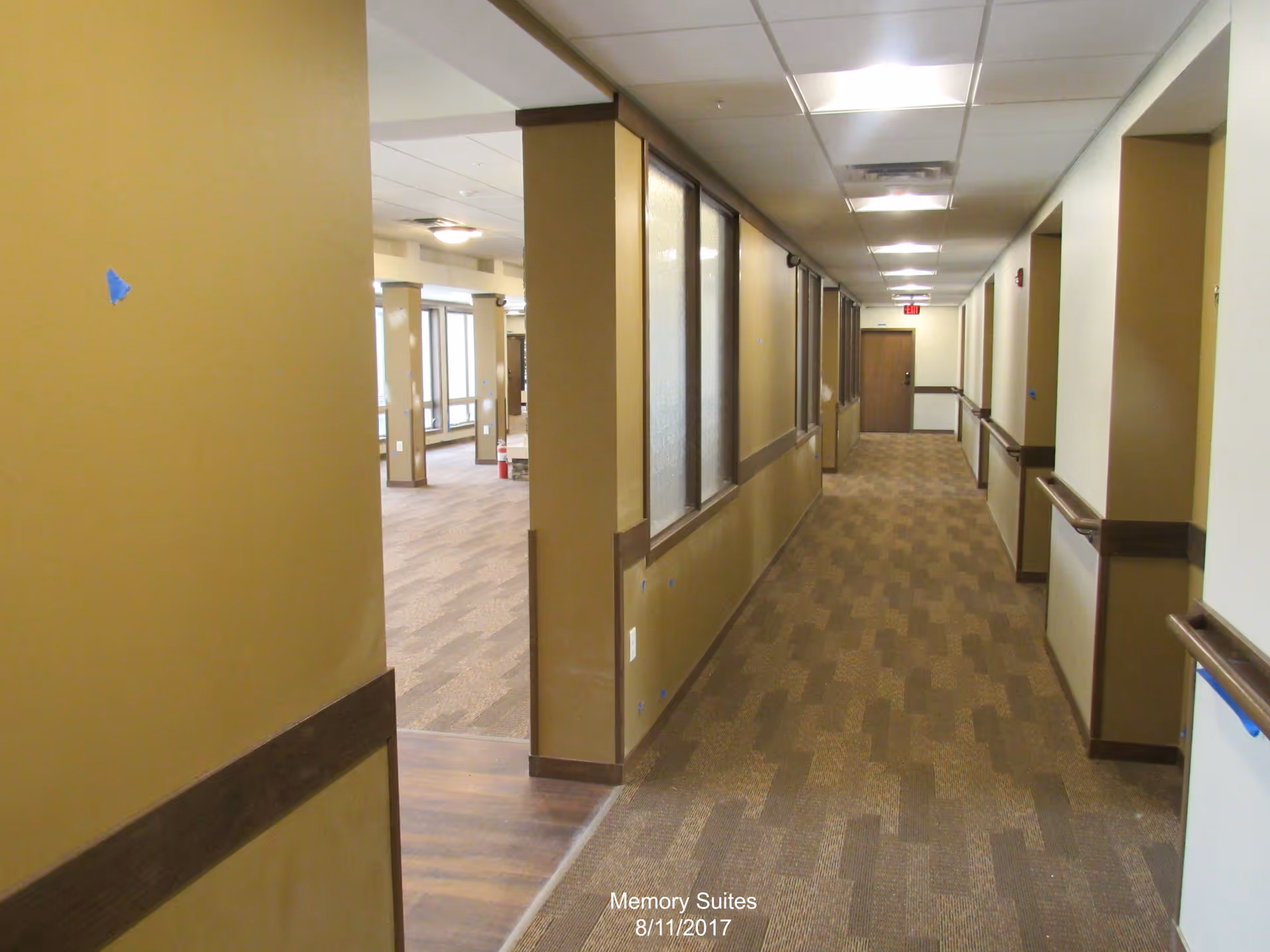 A long indoor hallway in a senior living facility with beige walls, brown handrails on both sides, and a patterned carpet floor. The hallway leads to a closed wooden door at the end. On the left side, there are windows and an open entrance to a larger room with large windows letting in natural light.