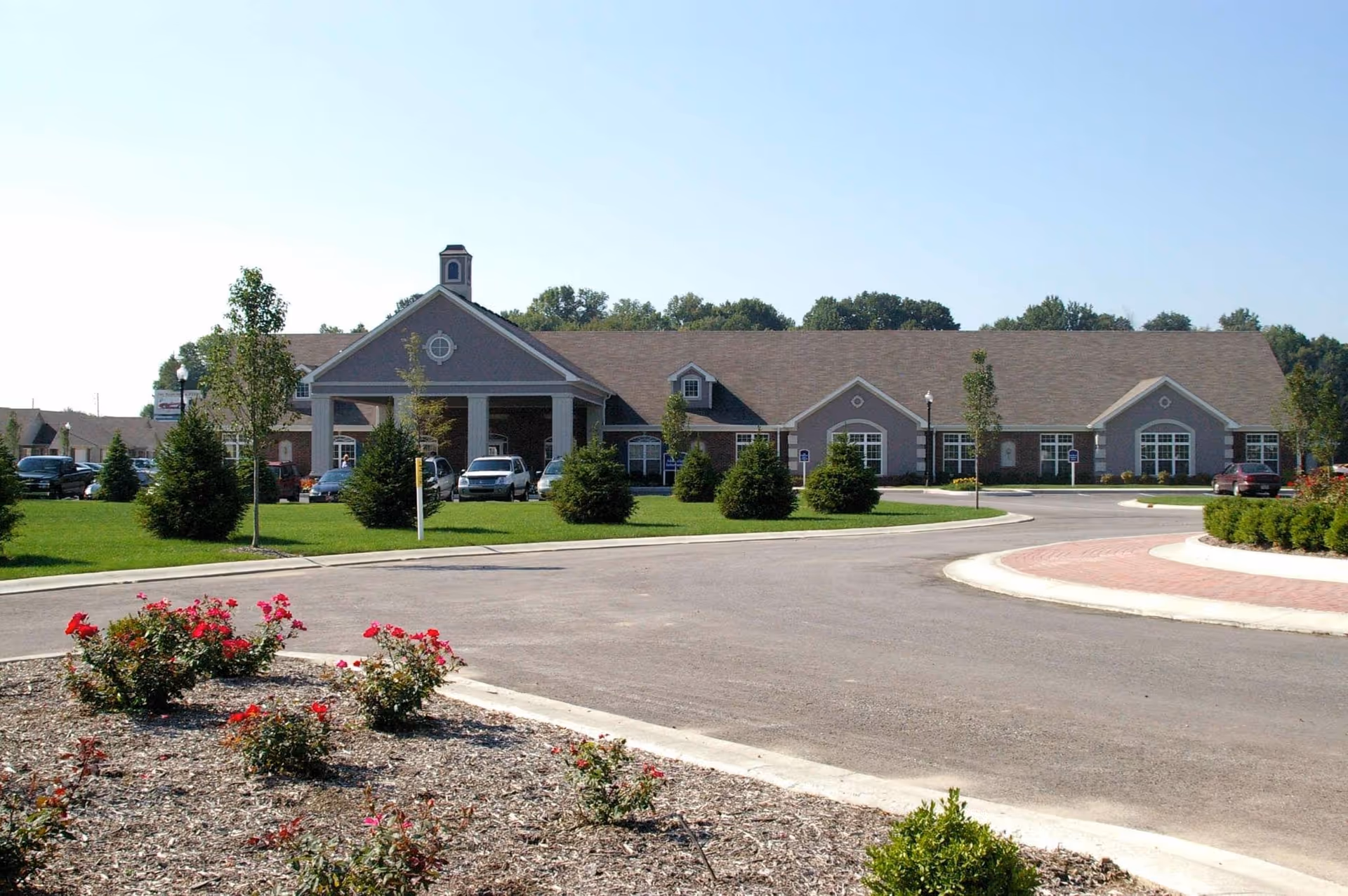 Exterior view of a single-story senior living facility building with a covered entrance, surrounded by a landscaped lawn with small trees and flower beds. Several cars are parked near the entrance under a clear blue sky.
