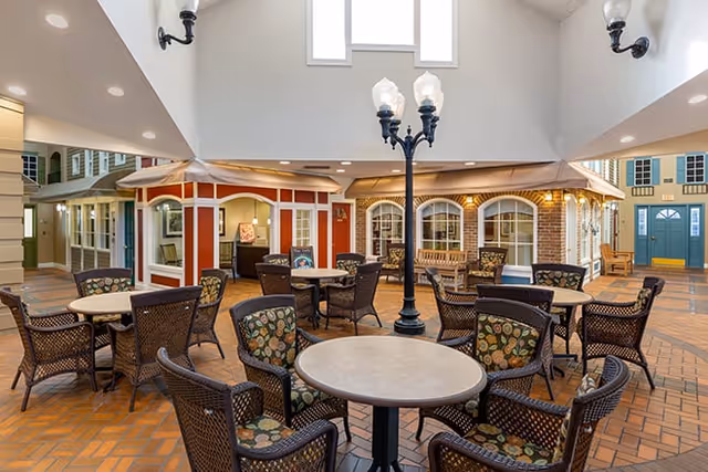Indoor common area with multiple round tables and wicker chairs with patterned cushions arranged around them. The space is designed to resemble an outdoor street scene with building facades, windows, and doors. There is a tall black street lamp in the center of the room and overhead lighting fixtures on the walls.