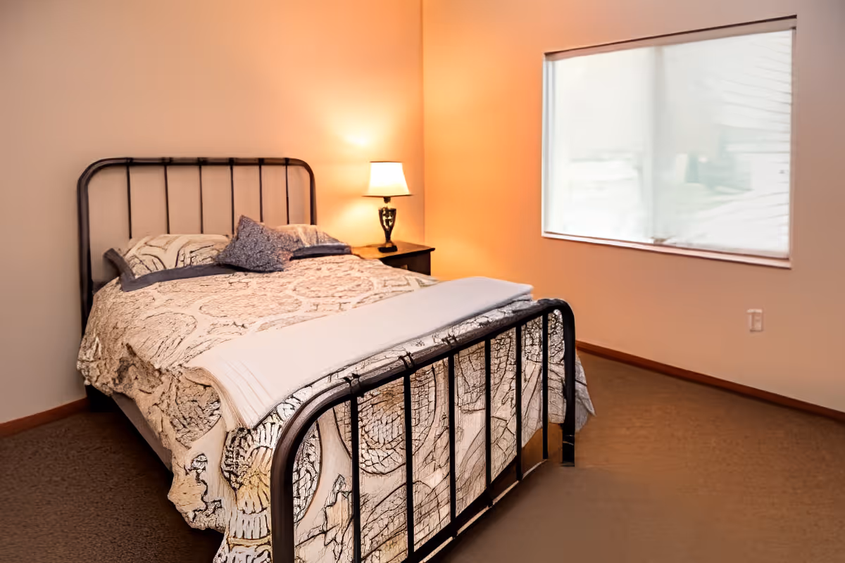 A cozy bedroom with a metal-framed bed covered in a patterned quilt and pillows. A bedside table with a lit lamp is next to the bed. There is a window with a white blind partially drawn, and the room has beige walls and carpeted floor.