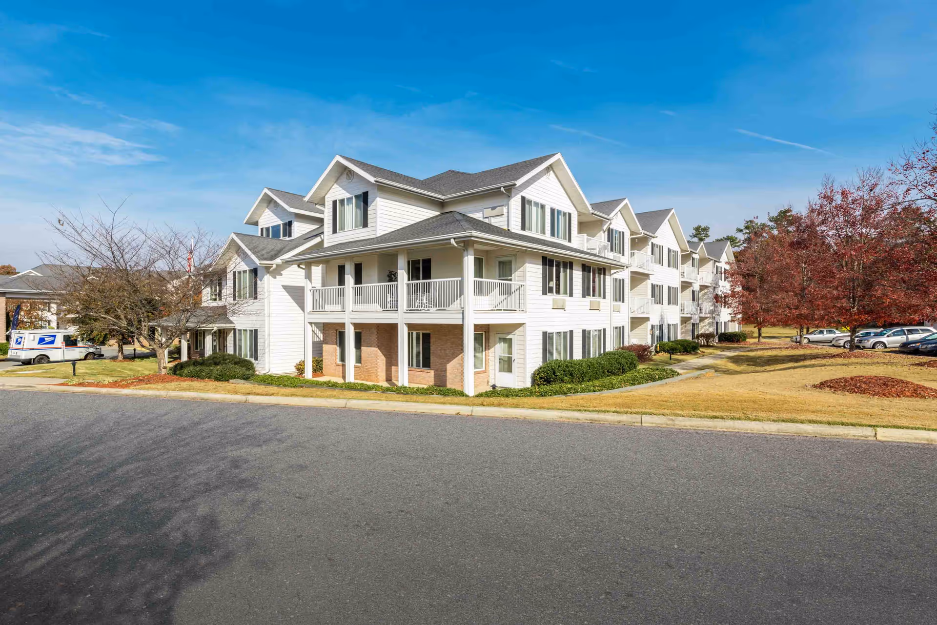 Large three-story white residential building with balconies and pitched roofs on a landscaped lawn and parking area under a blue sky.