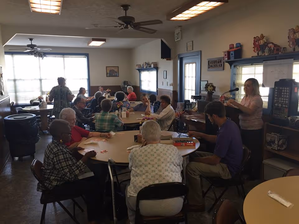 Seniors seated around tables in a communal activity/dining room playing games while a staff member speaks at a podium.