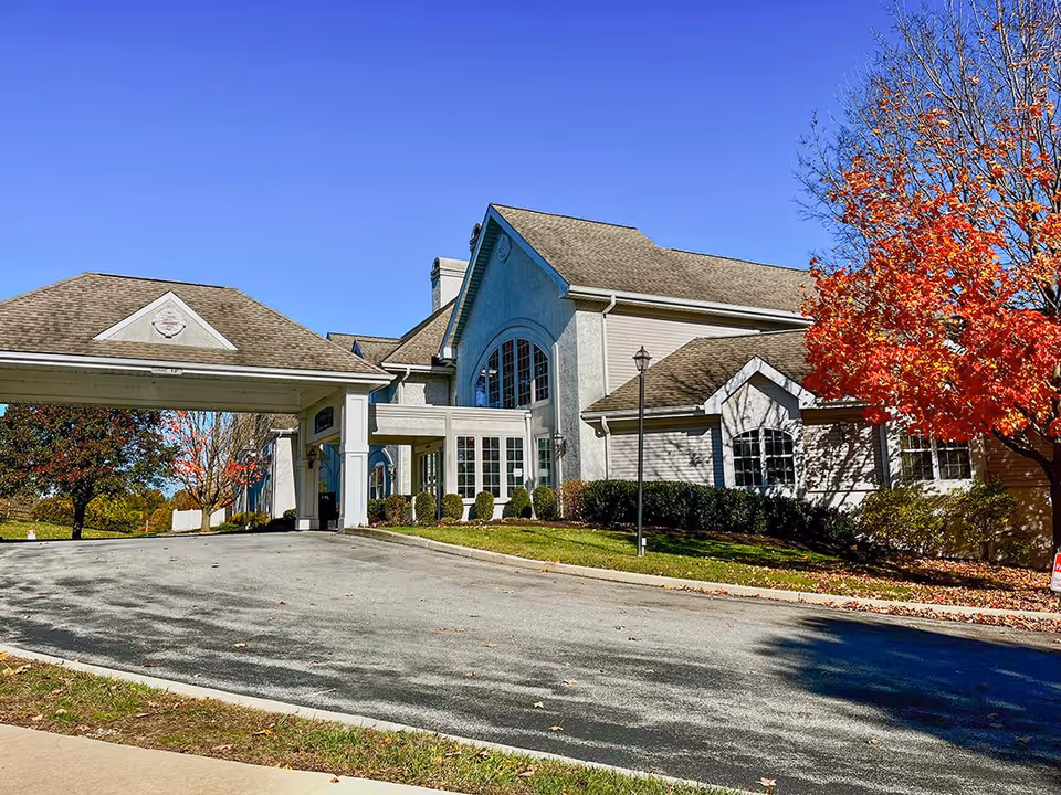 Exterior view of a senior living facility building with a covered entrance driveway, surrounded by trees with autumn foliage under a clear blue sky.
