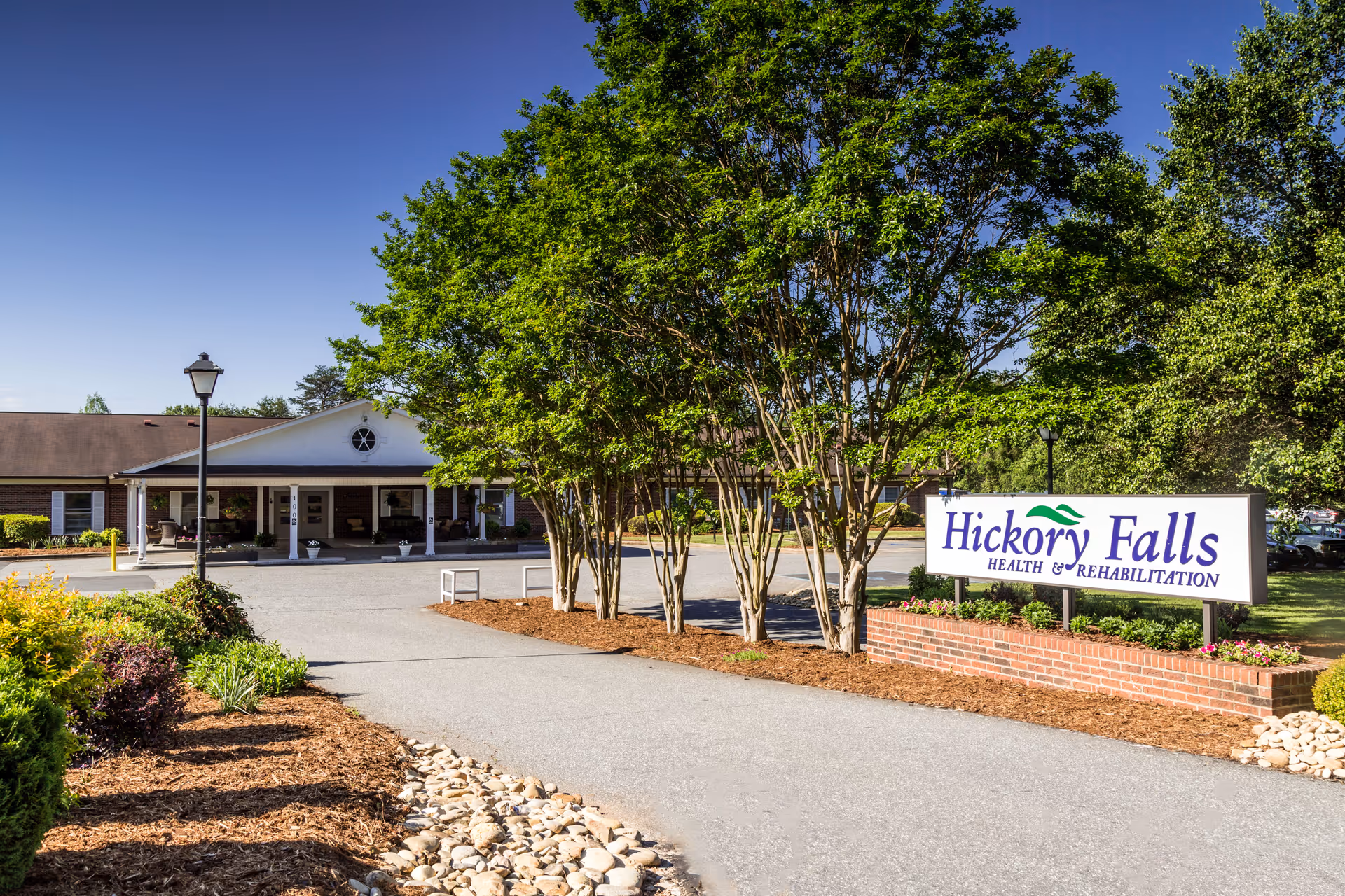 Entrance and sign for Hickory Falls Health & Rehabilitation with driveway, trees, and landscaped grounds in front of the building.