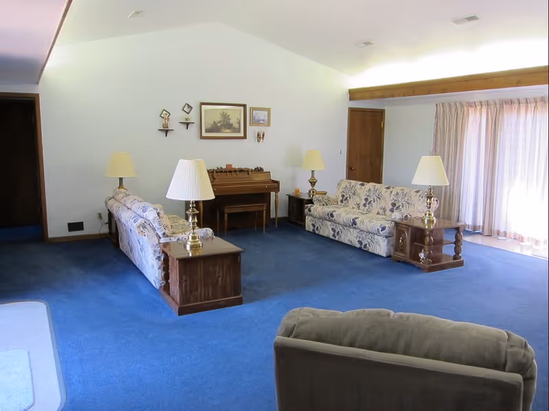 Spacious living room with blue carpet, floral sofas, wooden end tables with lamps, a piano against the wall, and large sliding curtains.