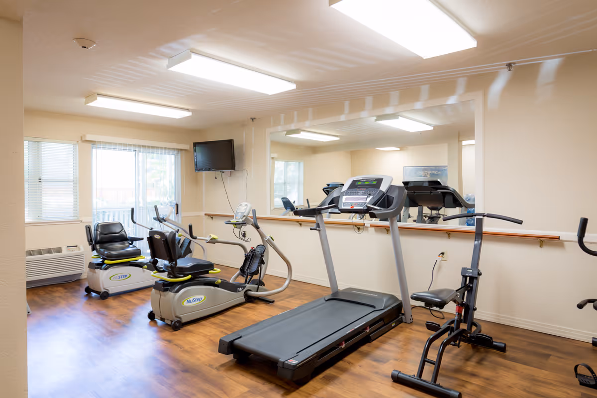 A small fitness room with exercise equipment including two NuStep recumbent cross trainers, a treadmill, and a rowing machine. The room has wood flooring, beige walls, a large mirror on one wall, fluorescent ceiling lights, a wall-mounted TV, and windows with blinds letting in natural light.