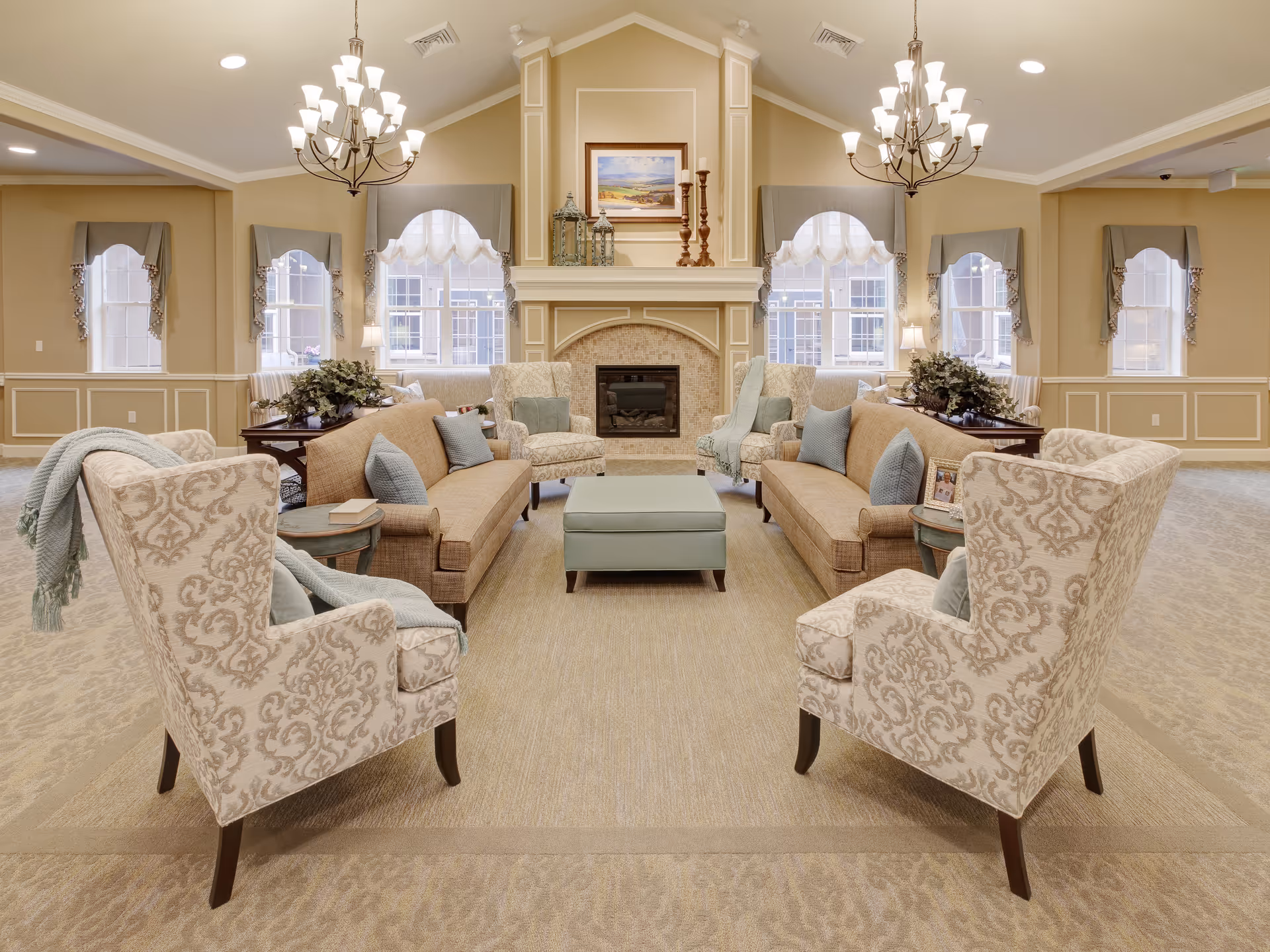 A spacious and elegant living room with two beige sofas facing each other, four patterned armchairs, a central ottoman, and a fireplace with decorative items and a painting above it. The room features large windows with draped curtains, two chandeliers, and soft beige walls and carpeting.