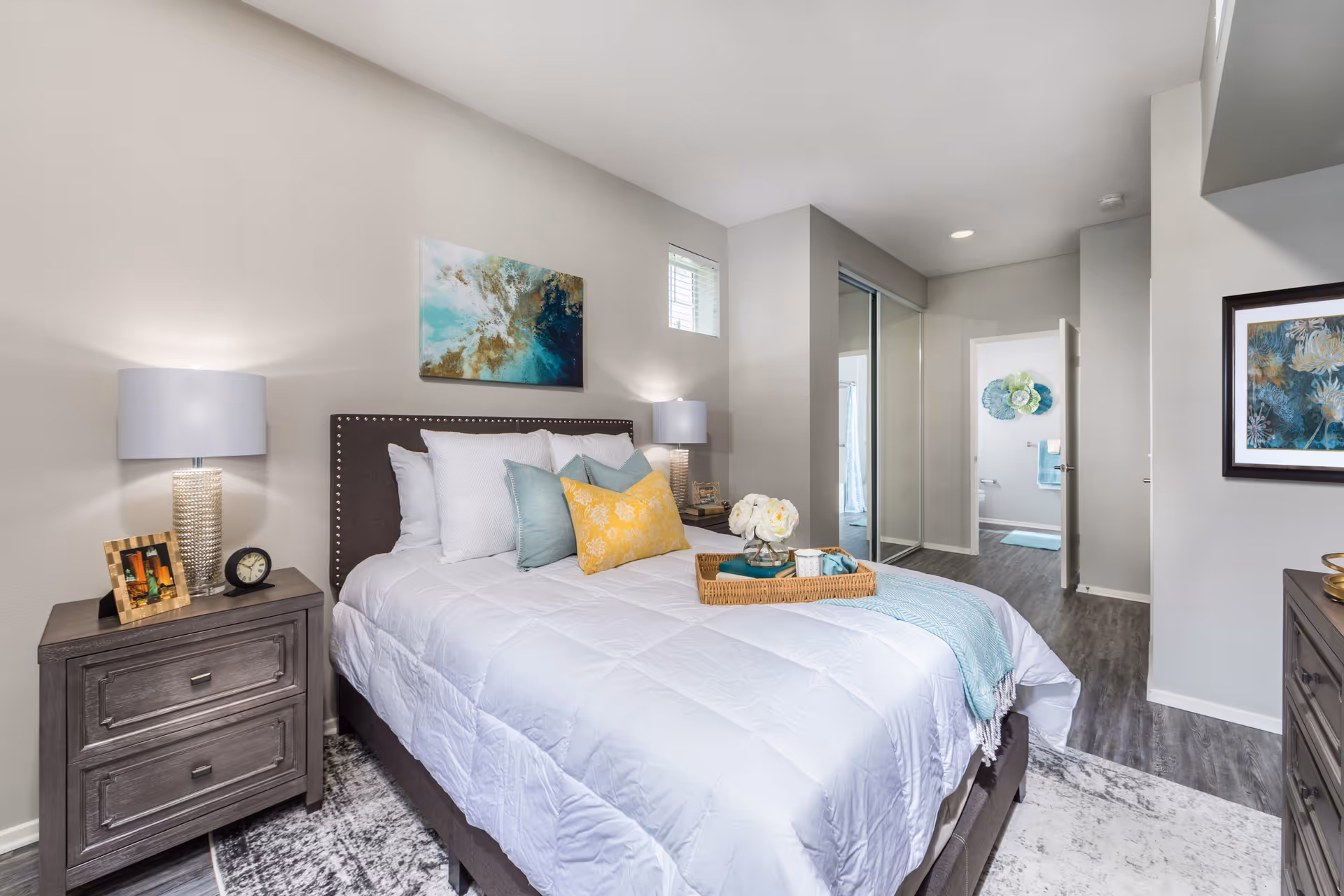 Bright modern bedroom with a made bed, bedside tables and lamps, artwork on the walls, and an en-suite bathroom visible through an open door.