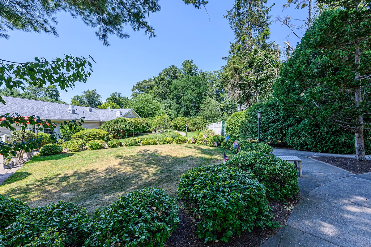 A landscaped outdoor garden area with trimmed bushes, a curved concrete pathway, and a bench. The garden is surrounded by tall trees and greenery under a clear blue sky. A building with a gray roof is partially visible on the left side.