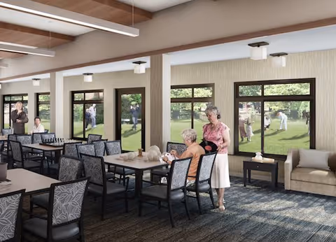 Communal dining/activity room with tables and chairs, seniors seated and standing near large windows overlooking a grassy courtyard.