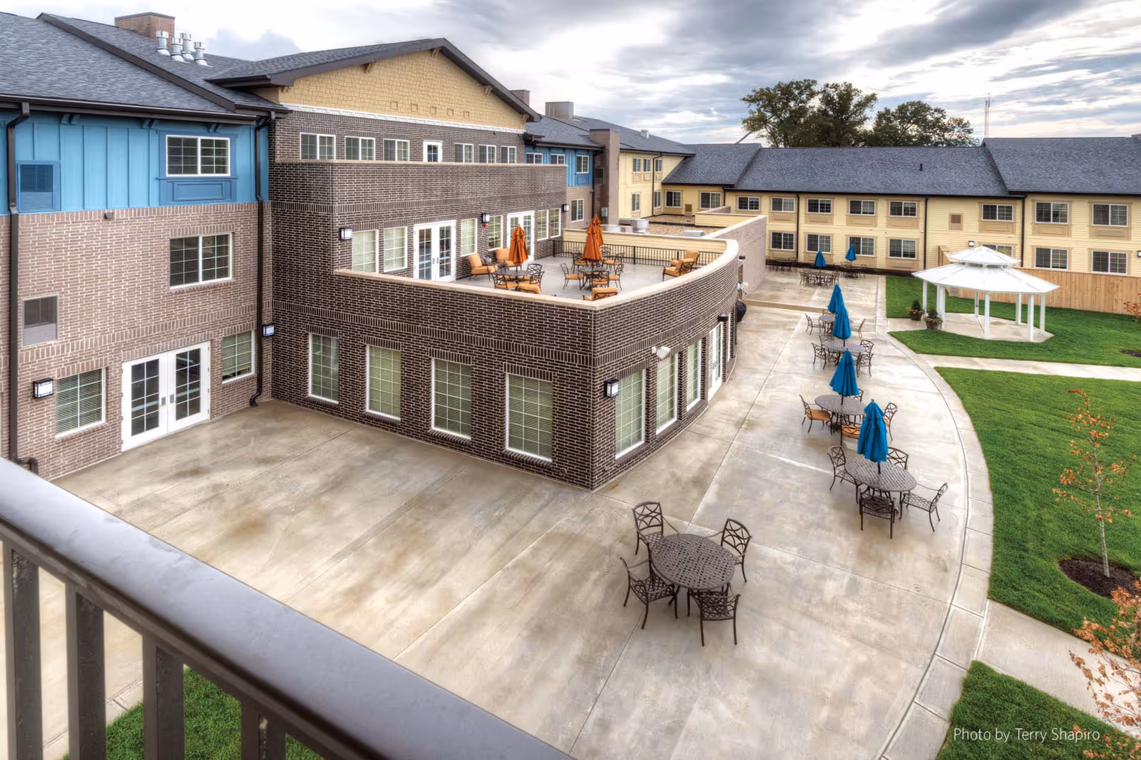 Outdoor patio area of a senior living facility with multiple round tables and chairs, some with blue umbrellas. The building has a brick exterior with a second-floor balcony featuring additional seating and orange umbrellas. There is a white gazebo on a grassy area adjacent to the patio.