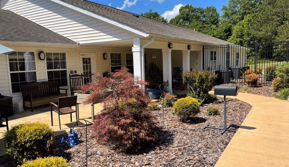 Outdoor patio area at Spring Arbor of Albemarle featuring a walkway, landscaped garden with bushes and a small tree, several wooden benches and chairs, and a building with white siding and multiple windows under a clear blue sky.
