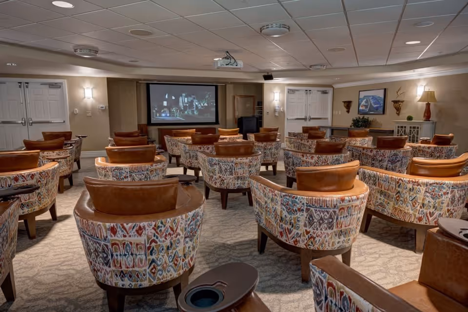 A cozy theater room with multiple rows of patterned armchairs featuring leather headrests, all facing a large screen at the front. The room has beige walls, carpeted floors, ceiling lights, and double doors on both sides of the screen. There is a lamp and framed pictures on the right wall.