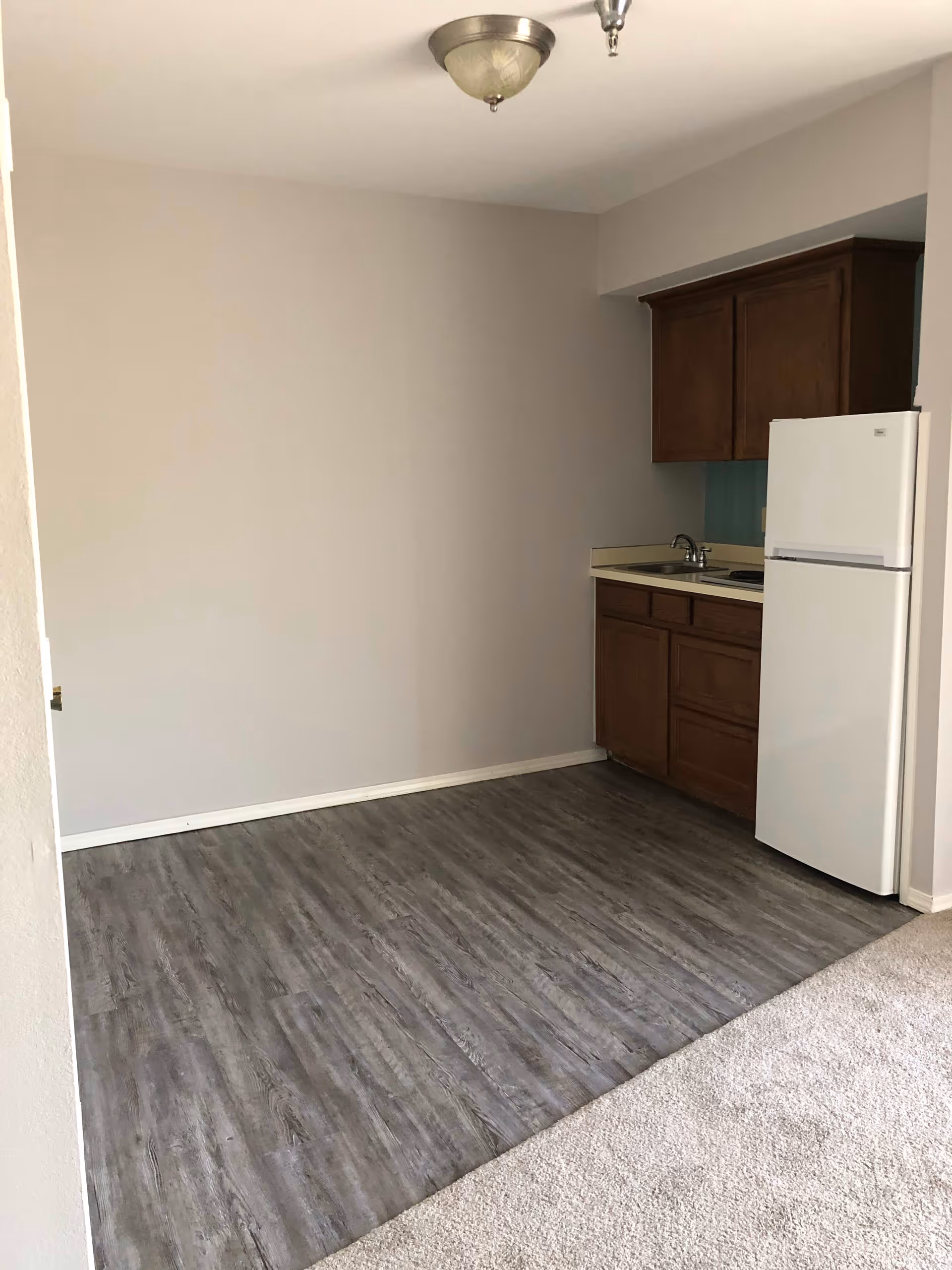 Small kitchen area with wooden cabinets, a white refrigerator, a sink, and a countertop. The floor has a wood-like finish, and the adjacent area is carpeted. The walls are plain and light-colored, with a ceiling light fixture visible.