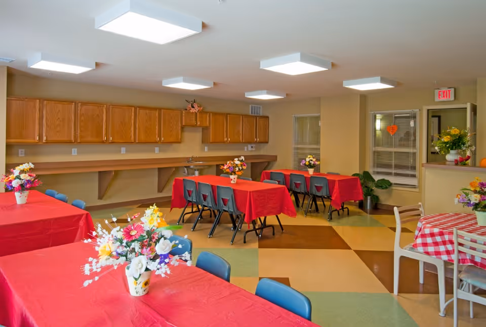 A bright dining room with several tables covered in red and red-and-white checkered tablecloths. Each table has a floral centerpiece. The room has wooden cabinets along one wall, a small sink, and a checkered floor with green, brown, and beige tiles. There are blue and white chairs around the tables, and a window with blinds and a door with an exit sign above it are visible in the background.