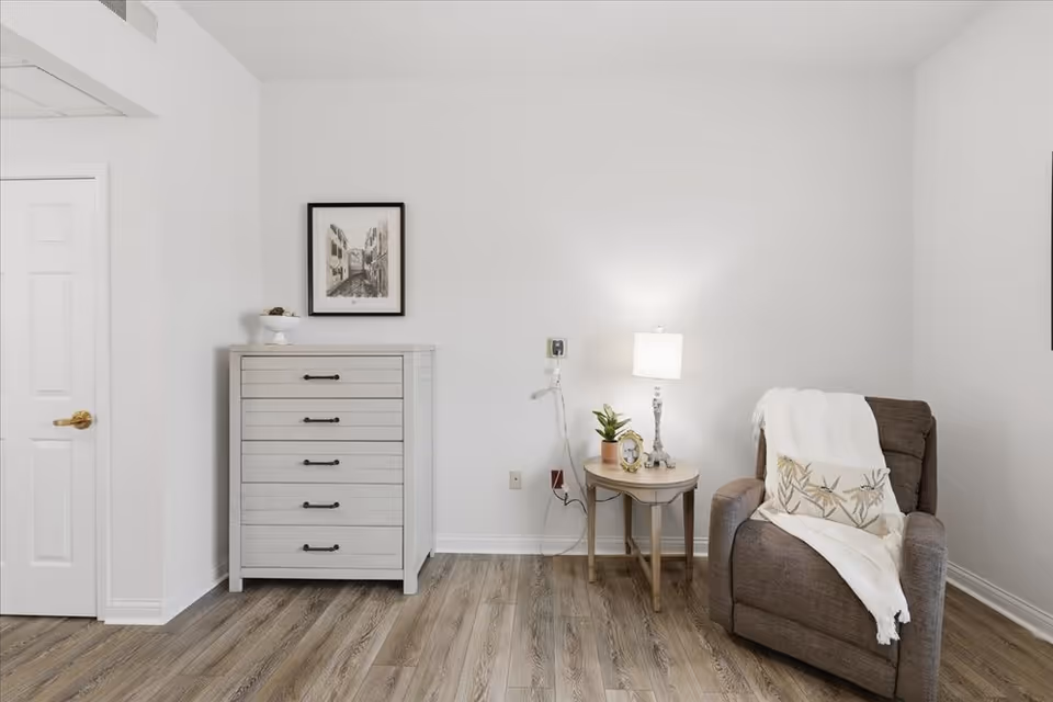 Bright minimalist living area with a white dresser, side table, lamp, and brown armchair