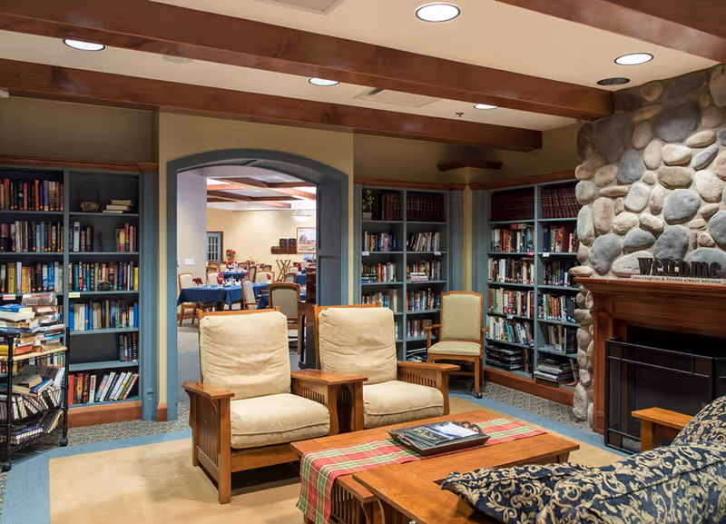 Cozy library/lounge with bookshelves, two upholstered armchairs, a wooden coffee table, and a stone fireplace, looking through an arch into a dining area.