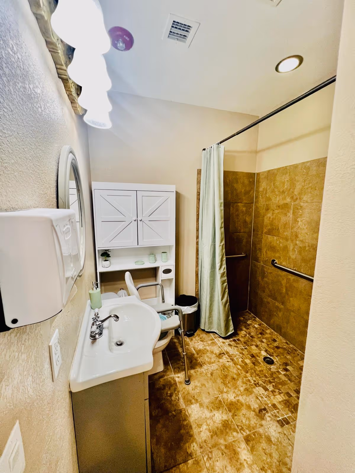 A small bathroom with beige tiled floor and walls, featuring a white sink with a silver faucet, a white cabinet with closed doors and shelves holding small items, a white chair with armrests, a trash can, and a walk-in shower area with a green shower curtain and grab bars on the walls.