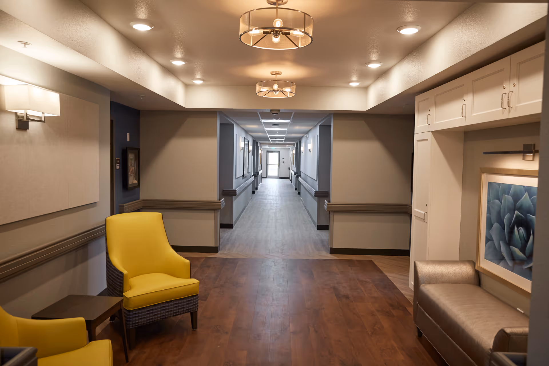 A well-lit hallway in a senior living facility with wood flooring and neutral-colored walls. The hallway features two yellow armchairs and a small table on the left, a beige cushioned bench on the right beneath a framed succulent artwork, and ceiling lights. The hallway extends into the distance with doors and handrails along the walls.