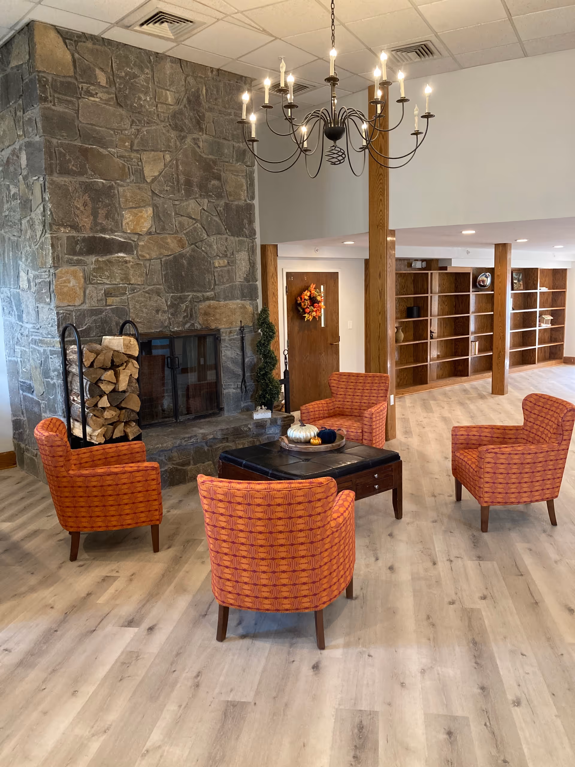 A cozy sitting area with four orange patterned armchairs arranged around a black ottoman in front of a large stone fireplace. There is a stack of firewood on a metal rack next to the fireplace. The room has light wood flooring, wooden pillars, a chandelier with lit bulbs, and built-in wooden shelves along the back wall. A door decorated with a fall wreath is visible in the background.