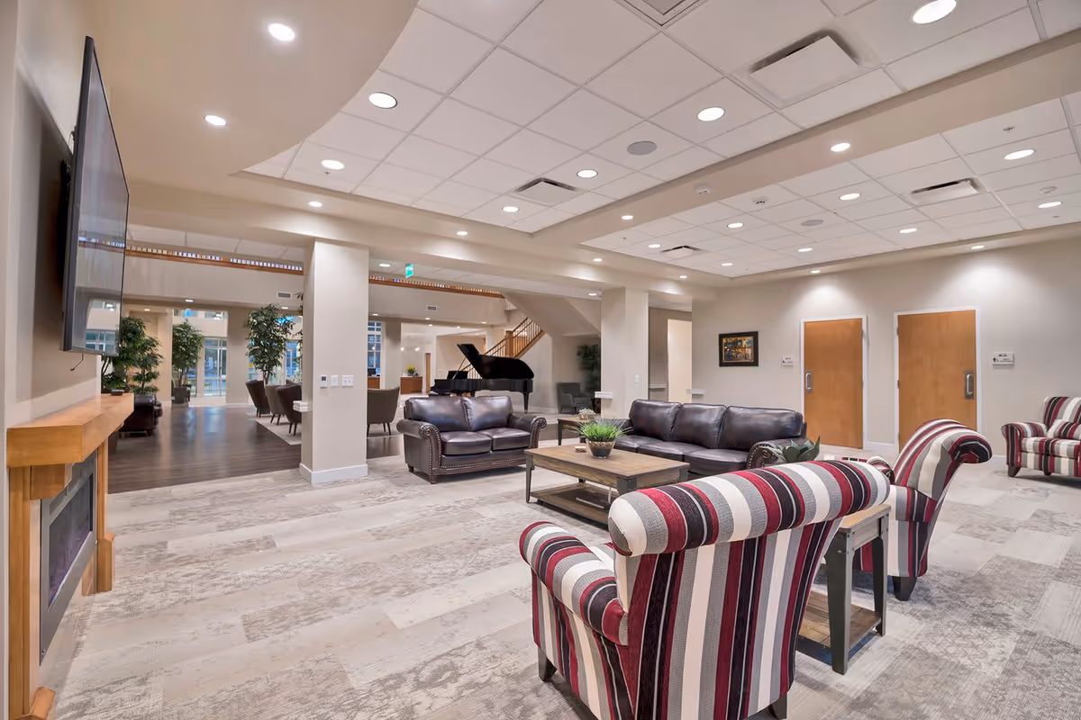 A spacious senior living facility common area with comfortable seating including striped armchairs and leather sofas arranged around a wooden coffee table. The room features a mounted flat-screen TV above a fireplace, recessed ceiling lights, and a grand piano in the background near large windows and plants.