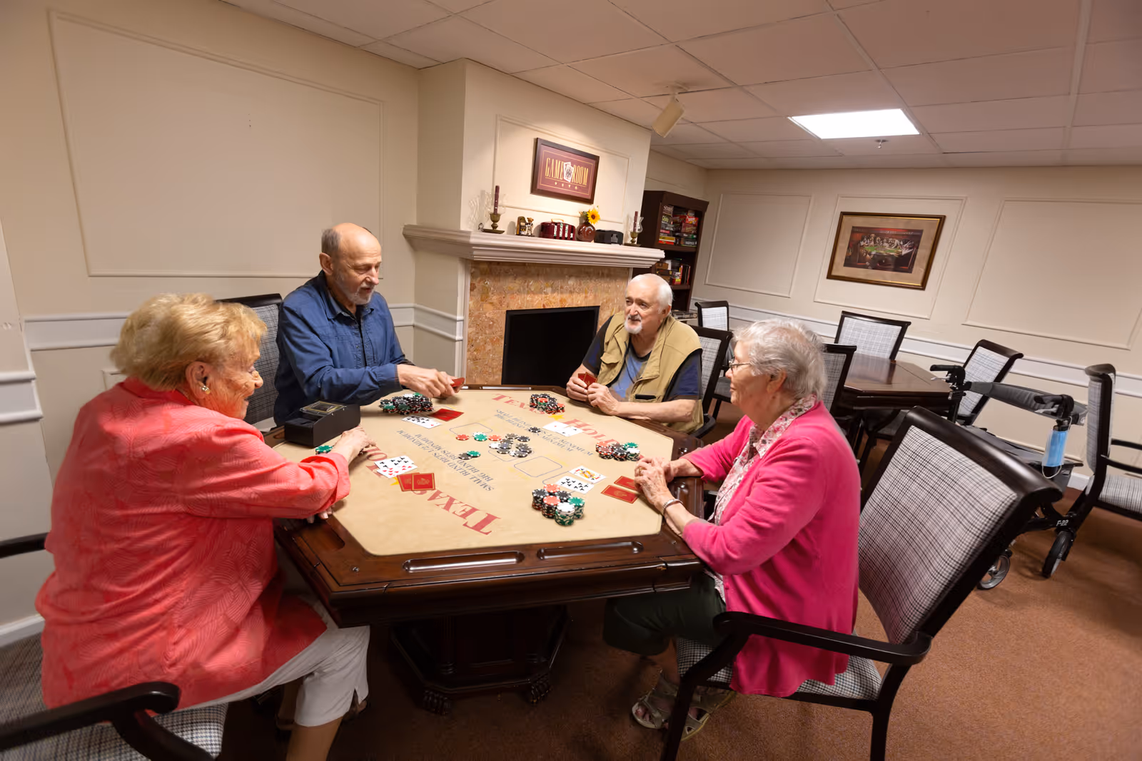 Four elderly people sitting around a card table playing poker in a cozy room with a fireplace, bookshelves, and framed artwork on the walls. The room has comfortable chairs and a walker is visible in the background.