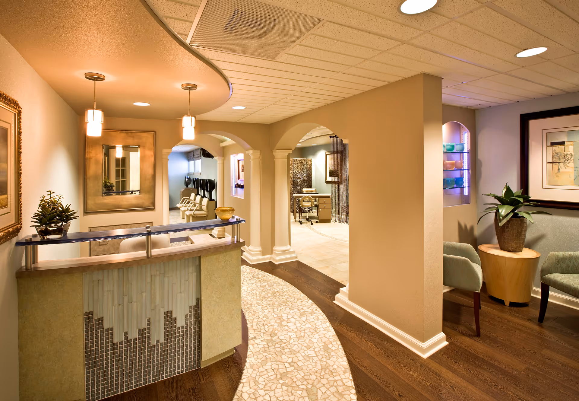 Reception area with a curved desk featuring mosaic tile design, two pendant lights hanging from the ceiling, a large mirror on the wall, and a small plant on the desk. The space has beige walls, wooden flooring with a curved tiled pathway, and arched doorways leading to other rooms. There is a seating area with two green chairs, a round wooden table with a potted plant, and framed artwork on the wall.