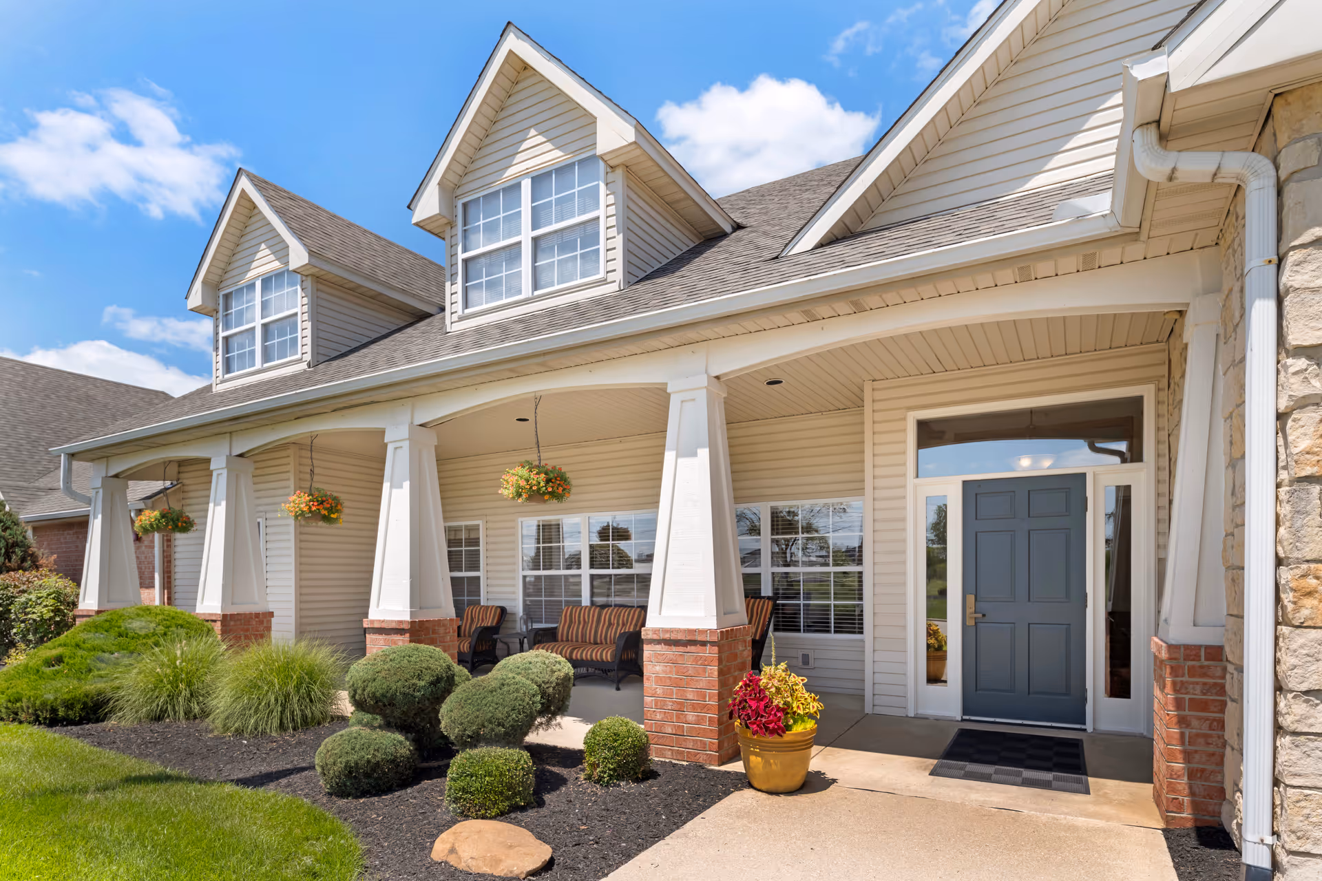 Front exterior view of a senior living facility with a covered porch featuring white columns with brick bases, hanging flower baskets, outdoor seating, and a blue front door under a clear blue sky.