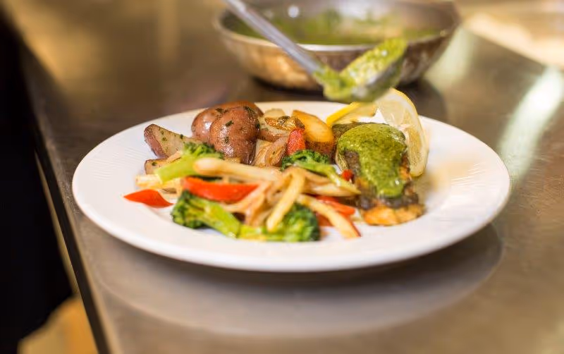 A white plate with a serving of cooked vegetables including broccoli, red bell peppers, and potatoes, alongside a piece of fish topped with green sauce and a lemon wedge. A spoon is seen adding more green sauce to the fish. The plate is on a stainless steel surface.