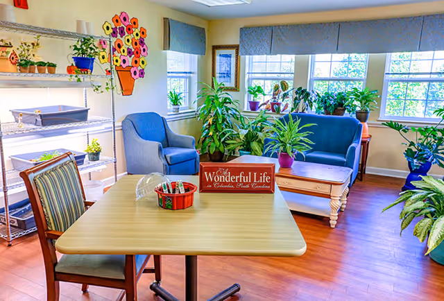 A bright and cozy common area with a wooden table and chairs in the foreground, a blue armchair and blue loveseat near windows with several potted plants on the windowsills and floor. A metal shelving unit with small plants and gardening supplies is on the left wall, and a colorful flower decoration is on the wall above the armchair. A sign on the table reads 'Wonderful Life in Columbia, South Carolina.'