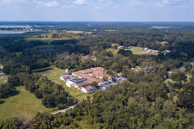 Aerial view of Stone Ledge Manor Senior Living facility surrounded by dense trees and greenery, with a few scattered buildings and open fields in the background under a partly cloudy sky.