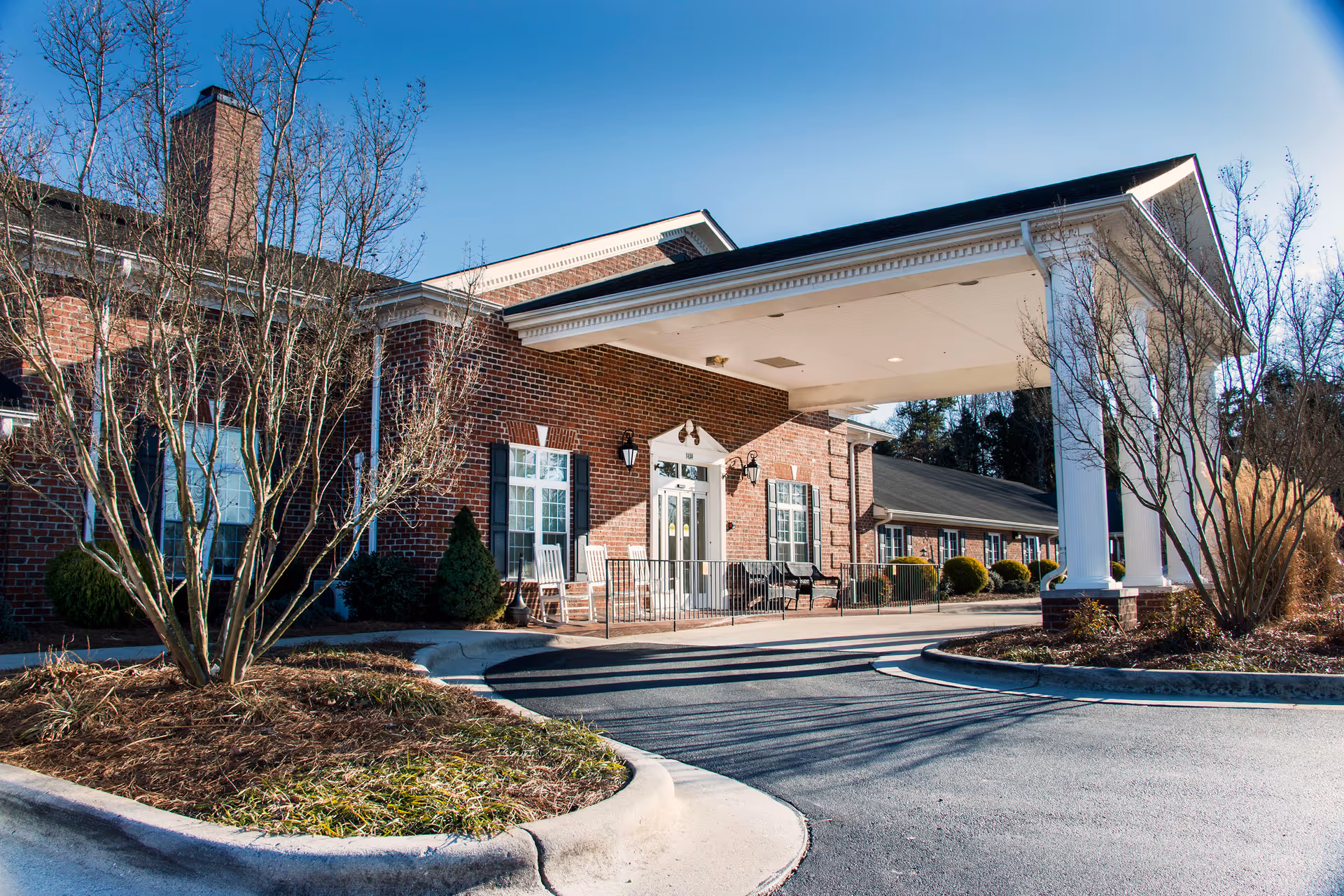 Front exterior view of a brick building with white columns supporting a covered entrance. There are leafless trees and shrubs around the driveway leading to the entrance. The sky is clear and blue.