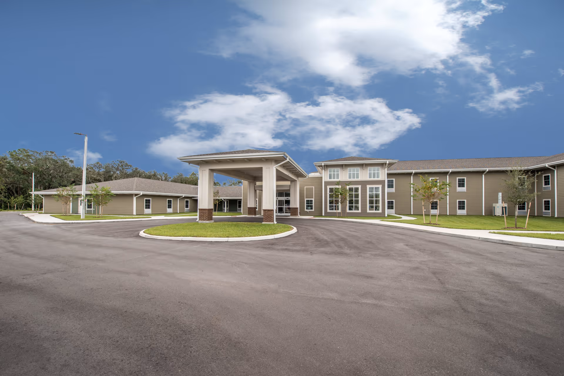 Exterior view of Twin Creeks Assisted Living & Memory Care facility showing a large, modern building with a covered entrance driveway, surrounded by a paved circular driveway and small landscaped areas with young trees under a partly cloudy blue sky.