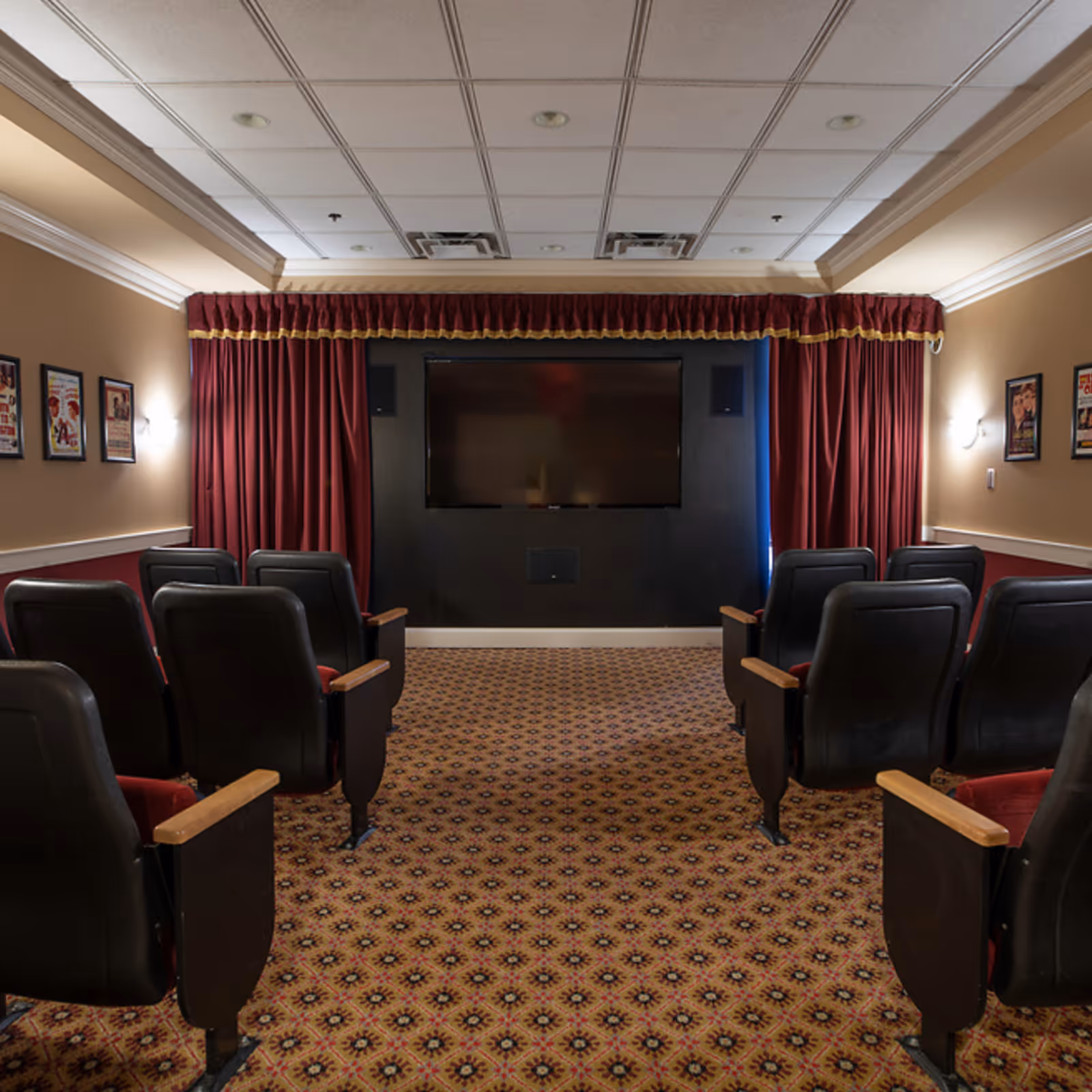 A small theater room with two rows of black cushioned seats with wooden armrests facing a large flat-screen TV mounted on a black wall. The room has patterned carpet, beige walls with red wainscoting, red curtains with gold trim framing the TV, and framed posters on the walls.