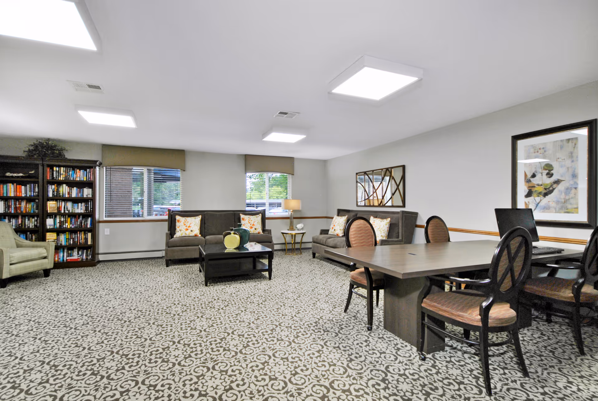 A well-lit common room with patterned carpet, two gray sofas with floral pillows, a coffee table with decorative vases, a bookshelf filled with books, a green armchair, a table with four chairs, a computer, and framed artwork on the walls.
