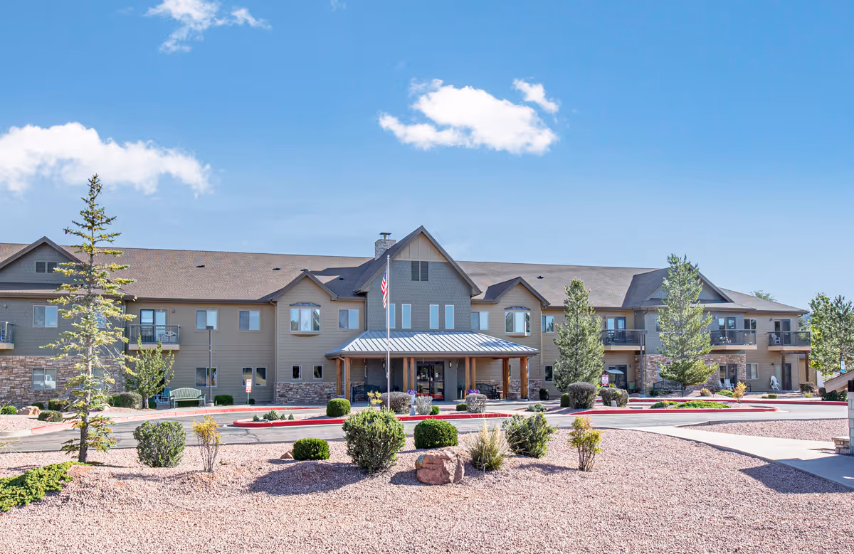 Exterior view of a two-story retirement living facility building with a covered entrance, surrounded by landscaped bushes, trees, and a gravel ground under a clear blue sky with a few clouds.