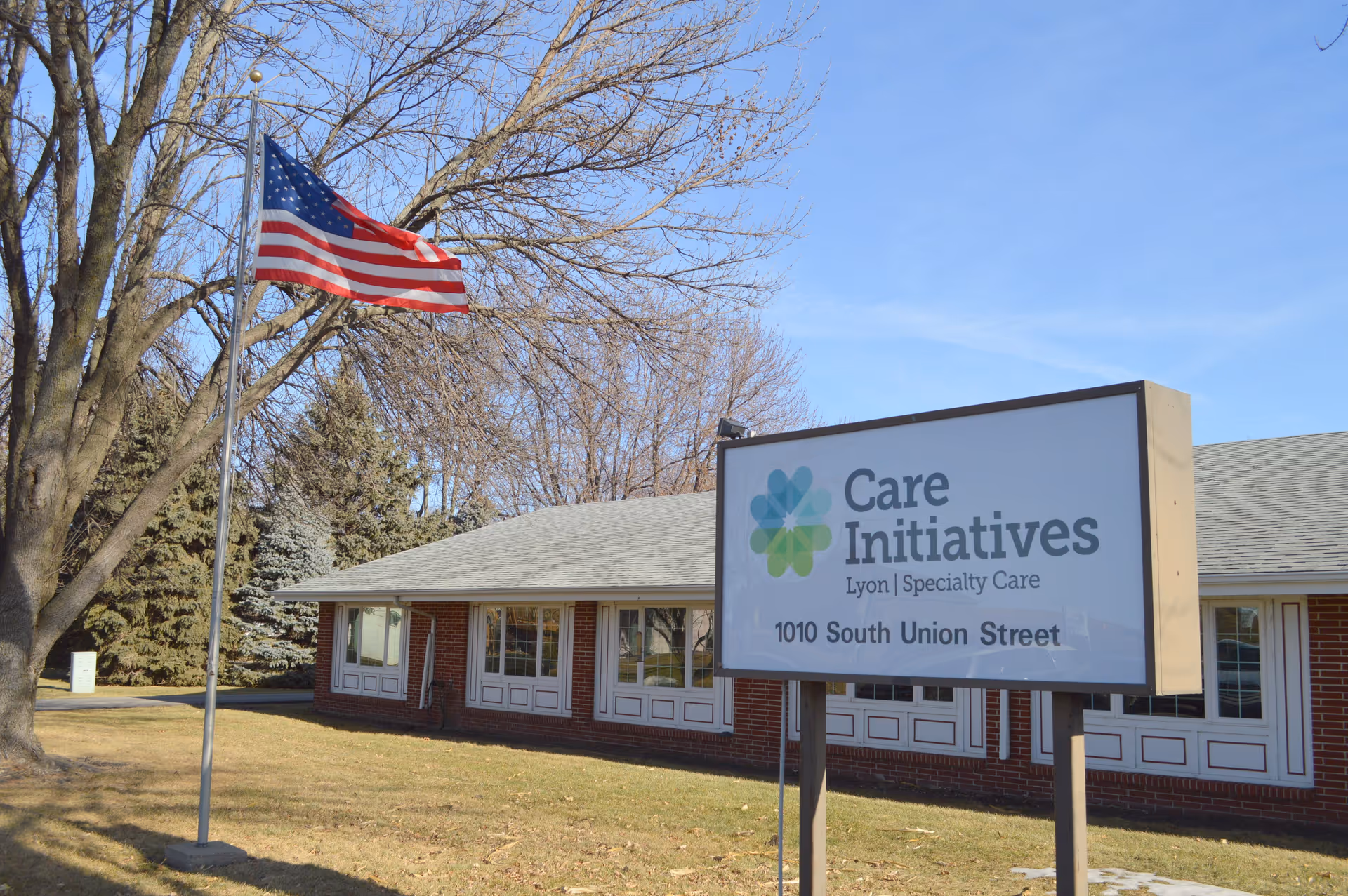 Exterior view of a single-story brick building with white framed windows, a large sign in front reading 'Care Initiatives Lyon Specialty Care 1010 South Union Street,' and an American flag on a flagpole nearby. Trees without leaves are visible in the background under a clear blue sky.