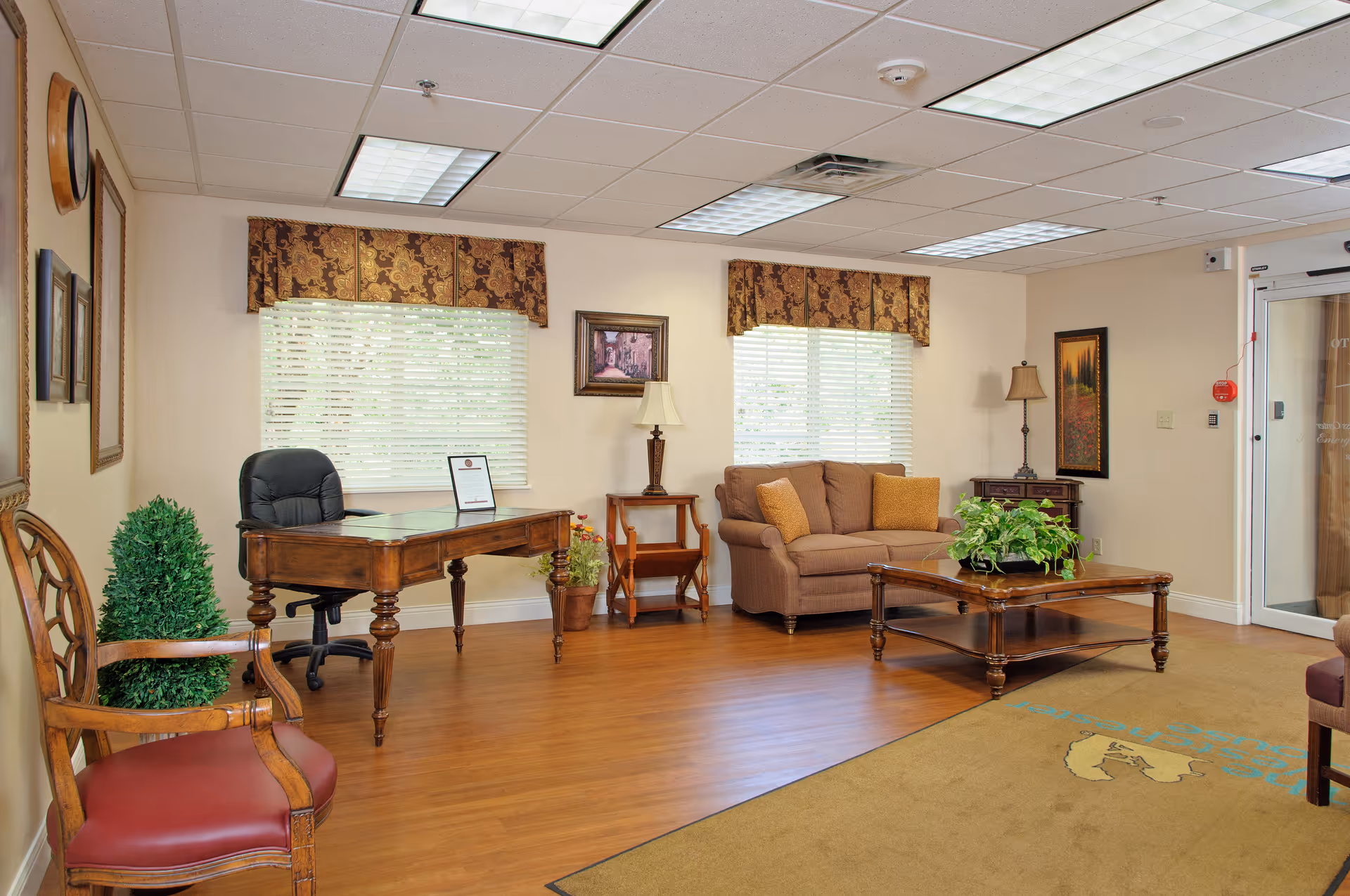 A well-lit interior room with wooden flooring featuring a wooden desk with a black office chair, a brown upholstered sofa with two cushions, a wooden coffee table with a green plant, and several framed pictures on cream-colored walls. The room has two windows with patterned valances and white blinds, a couple of table lamps, and a glass door on the right side.