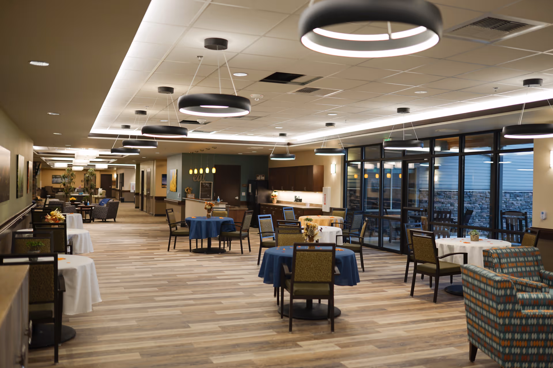 Interior view of a spacious dining area in a senior living facility with several round tables covered with blue and white tablecloths, chairs arranged around them, modern circular ceiling lights, large windows on one side, and a kitchenette area in the background.