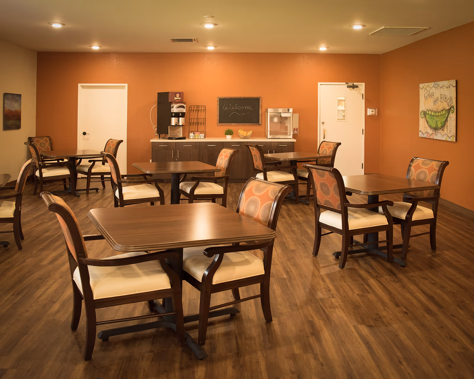 Dining room with multiple wooden tables and cushioned chairs, a coffee station and warm orange walls.