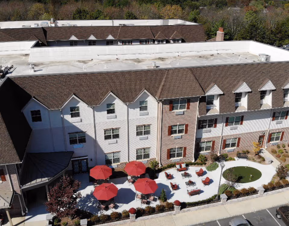 Aerial view of Avalon Assisted Living at Hillsborough showing a multi-story building with brown shingled roofs and white and brick exterior walls. In front of the building is a patio area with several red umbrellas, outdoor seating, and a small circular putting green. There are landscaped bushes and trees around the patio and a parking lot visible at the bottom of the image.