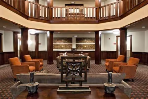 Interior senior-living lounge with red chairs, sofa, wood columns, and upper balcony.