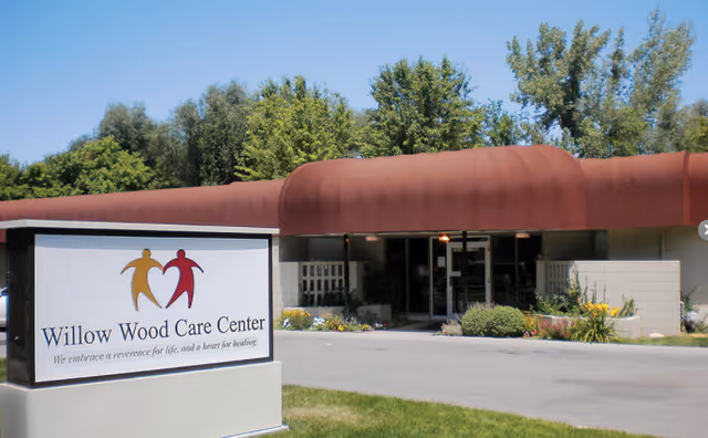 Exterior view of Willow Wood Care Center building with a brown curved awning over the entrance, surrounded by greenery and flowers. A large sign in front displays the facility name and logo with two stylized human figures holding hands.