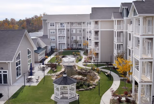 Outdoor courtyard area of a senior living facility featuring a white gazebo, a small pond with a stone border, walking paths, benches, and multi-story residential buildings surrounding the courtyard. Trees with autumn foliage and outdoor seating areas are also visible.