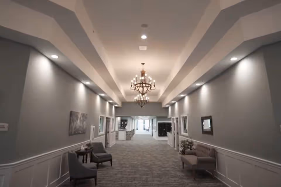 A long, well-lit hallway with gray walls and carpeted floor in a senior living facility. The hallway features recessed lighting along the ceiling edges and two chandeliers hanging from the ceiling. There are chairs and small tables placed along the walls, with framed artwork and mirrors decorating the space.