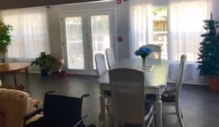 Sunlit dining area with a table and chairs, a wheelchair, potted plants, and glass doors with curtains.