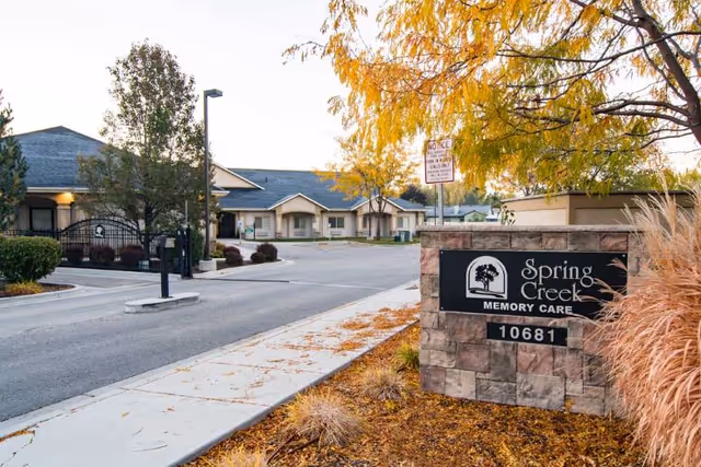 Exterior view of Edgewood Spring Creek Memory Care facility with a stone sign displaying the name and address 10681, a paved driveway, sidewalk, trees with autumn foliage, and single-story buildings in the background.