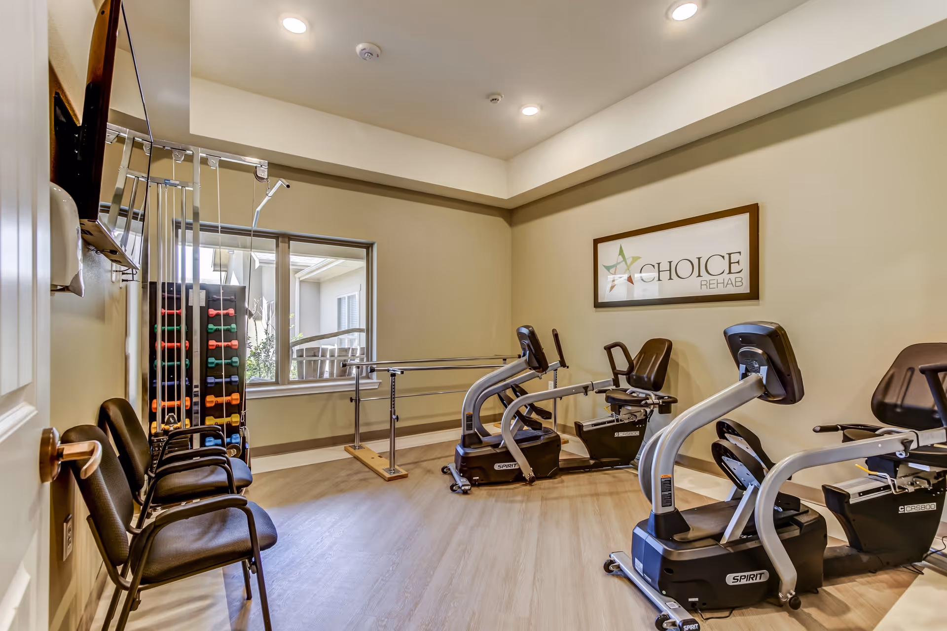 A small rehabilitation exercise room with two recumbent exercise bikes, a set of colorful dumbbells on a rack, parallel bars for walking support, and three black chairs lined up against the wall. A framed sign on the wall reads 'Choice Rehab'. There is a window showing an outdoor view and the room has recessed ceiling lights.