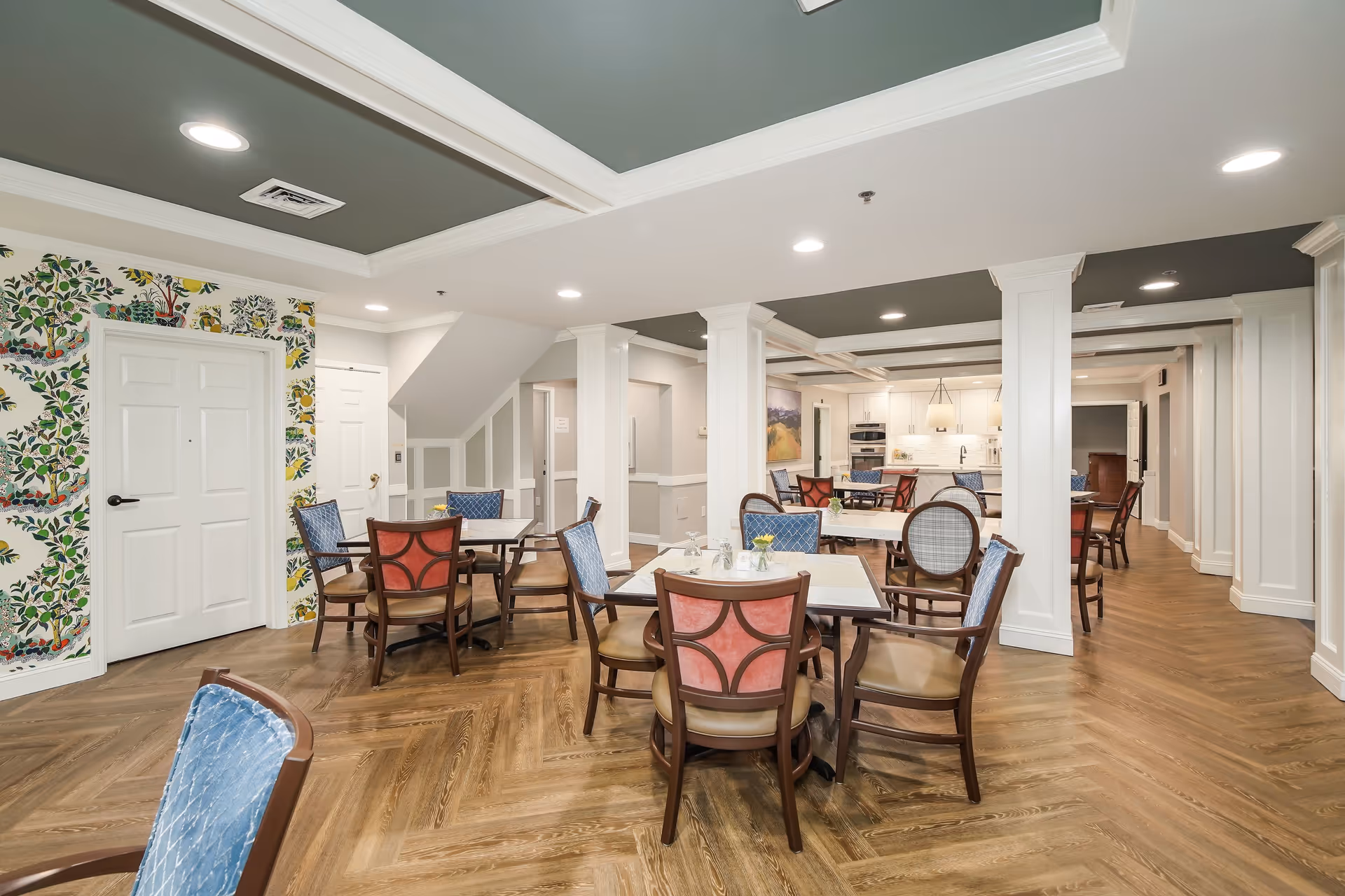 A bright and spacious dining area with multiple tables and chairs arranged neatly. The room features a patterned wallpaper with leafy designs on one wall, white doors, and white columns supporting the ceiling. The ceiling has recessed lighting and a coffered design painted in a muted green color. The floor is covered with wood-patterned flooring arranged in a herringbone style. In the background, a kitchen area with white cabinetry and modern appliances is visible.