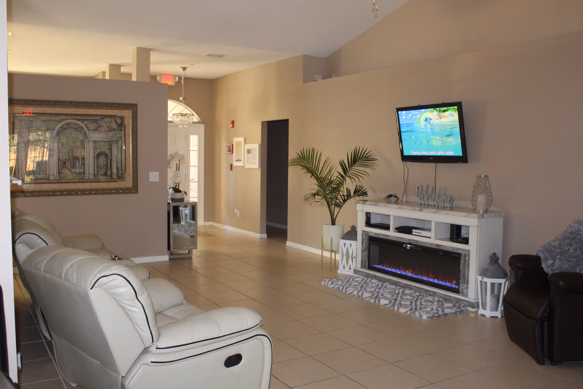 A living room area with beige walls and tiled floor. There are two light-colored recliners on the left and a dark armchair on the right. A white entertainment console with an electric fireplace is against the wall, with a flat-screen TV mounted above it. A potted plant and decorative lanterns are placed near the console. A framed painting hangs on the left wall, and a hallway with a door is visible in the background.